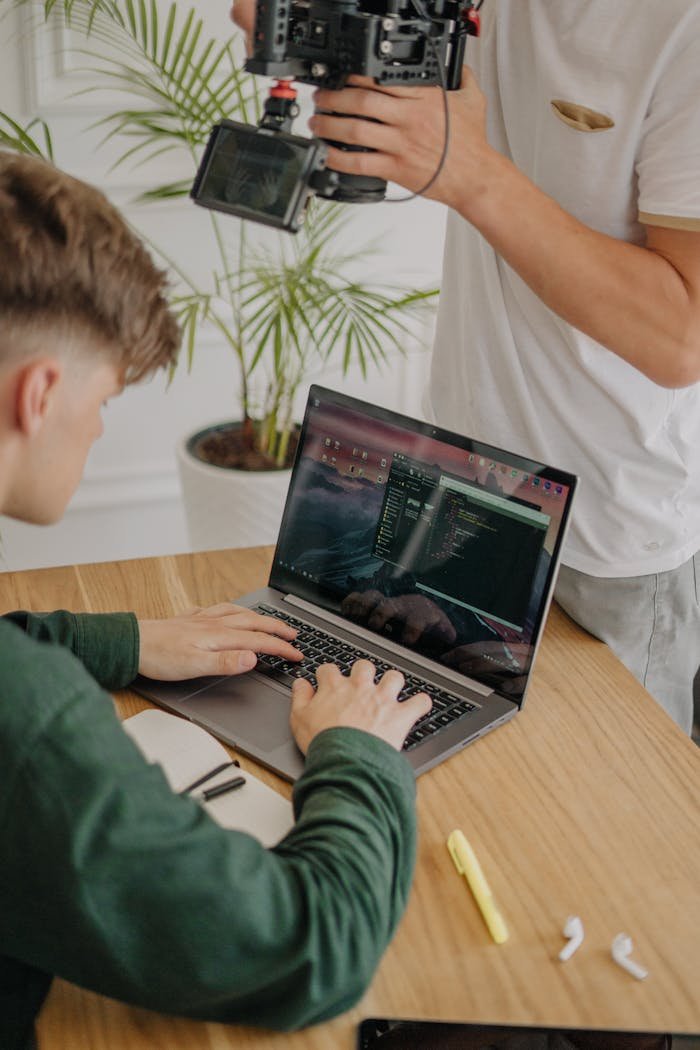 A young videographer records while a boy types code on a laptop at a wooden desk indoors.