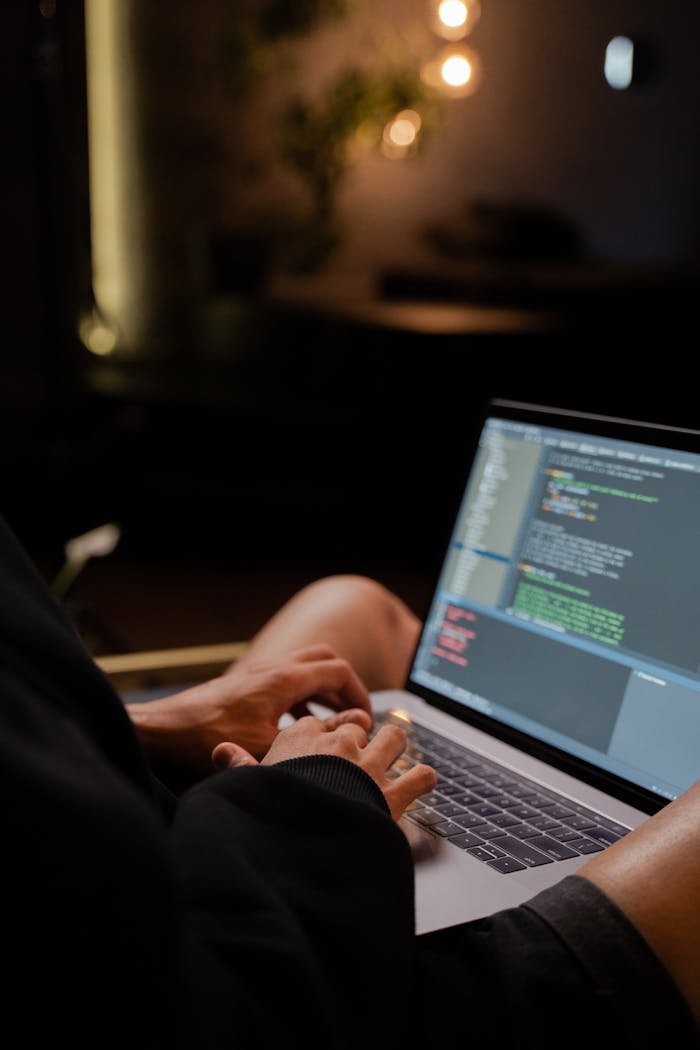 Focused hands typing code on a laptop in a dimly lit room, showcasing programming activities.